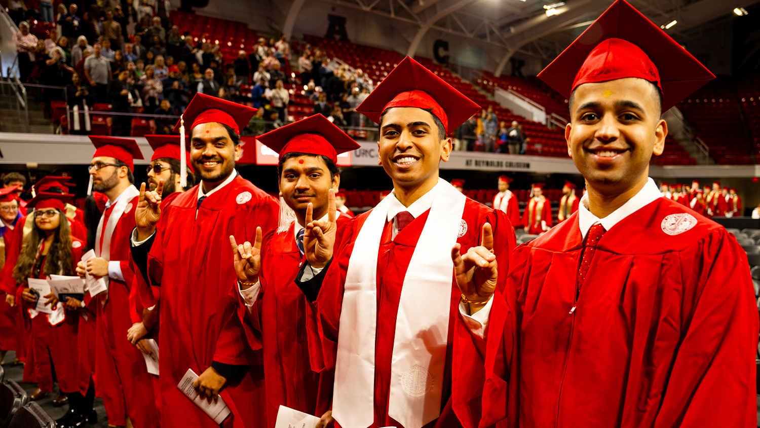 A group of smiling graduates in red caps and gowns with white stoles are standing in an arena. A large audience is visible in the background.