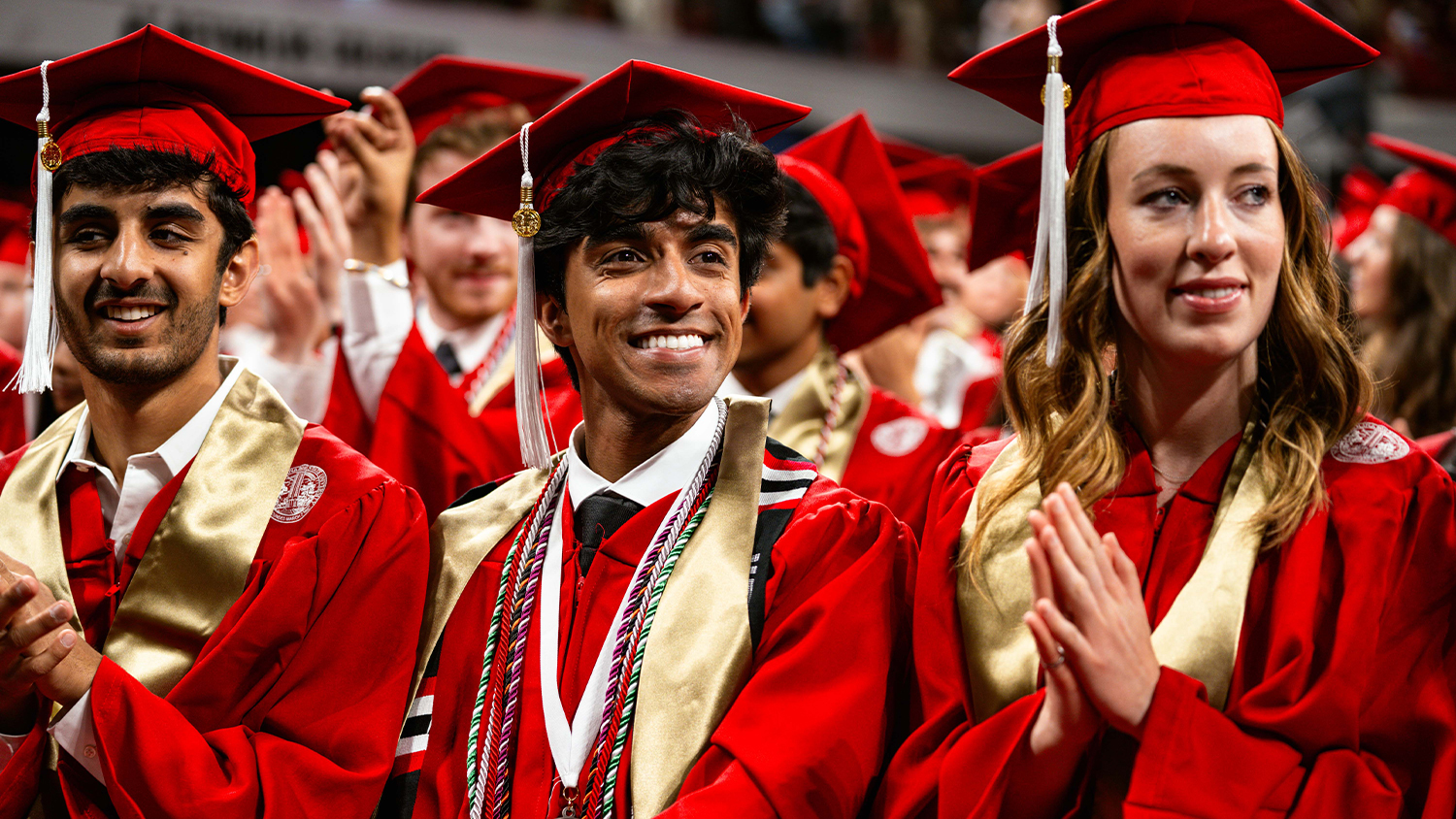 A group of smiling graduates from North Carolina State University, are captured in a close-up shot during a graduation ceremony. They are all wearing bright red caps and gowns. From left to right: Leftmost graduate: Appears to be a man with a beard and dark hair, smiling and looking towards the camera. He has a gold stole around his neck. Center graduate: Appears to be a young man with dark, curly hair, looking slightly to his right with a wide smile. He is adorned with multiple honor cords and a gold stole. Rightmost graduate: Appears to be a woman with long, light brown hair, looking slightly to her left with a gentle smile. She has a gold stole draped over her shoulders. In the background, other graduates in similar attire are visible, some clapping, adding to the festive atmosphere of the graduation.