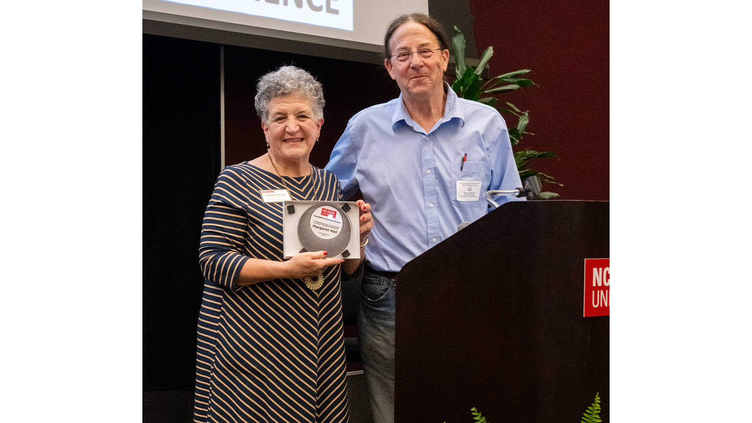 A man and a woman are standing next to a podium, both smiling at the camera. The woman, on the left, has short, curly grey hair and is wearing a dark blue and yellow striped dress. She is holding a plaque or award with a circular design. The man, on the right, has longer dark hair pulled back and is wearing a light blue collared shirt and jeans.