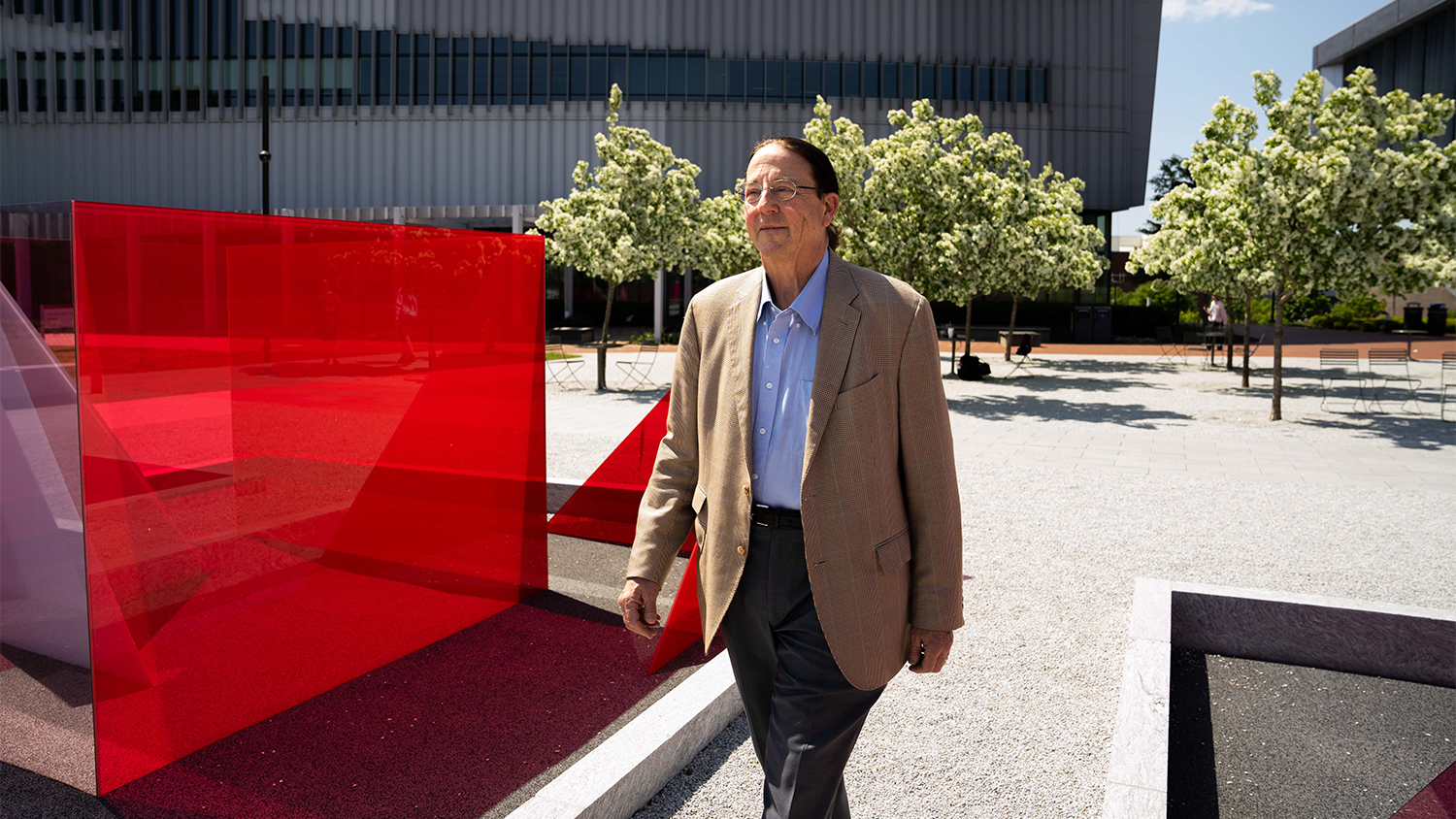 Gregg Rothermel, a man with glasses and a light brown suit jacket walks on a path between a bright red art installation and a few small trees with white blossoms. The man is looking slightly to his left and has a calm expression. In the background are more trees and a modern building.