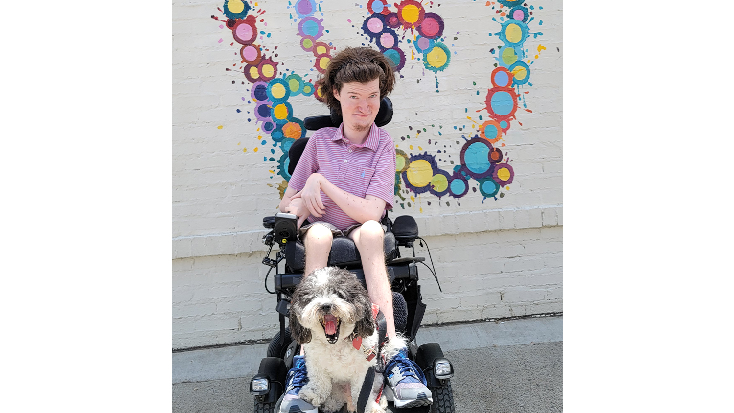Michael 'Gray' Dougherty in a wheelchair with a dog on his lap sits in front of a colorful mural.