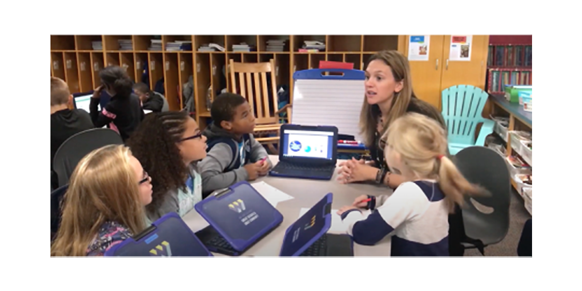 A female teacher sits at a small table with four young students, appearing to be in an elementary school classroom. She is gesturing while talking to them. On the table, there are two laptops open in front of the students. One of the laptops is displaying a screen with a pie chart and other data visualizations, likely related to an educational topic. The students are attentively looking at the teacher or the screen. In the background, there are shelves filled with books and cubbies, and other students are working on computers.