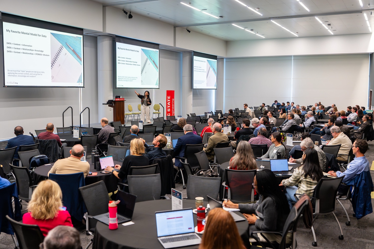 A photo of dozens of attendees in a well lit conference hall, looking at a speaker on stage.