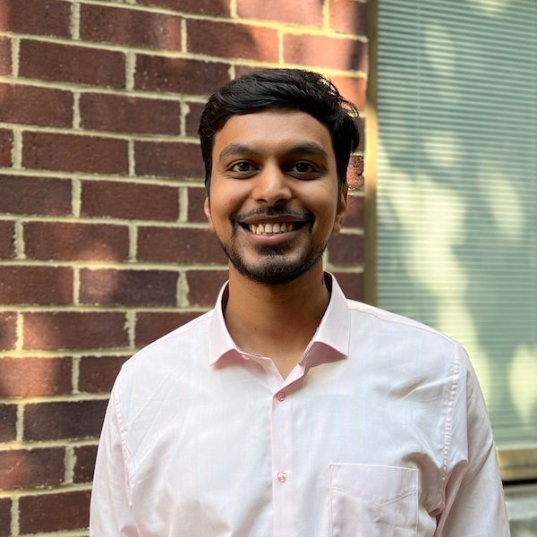 A man with dark hair and a short beard, wearing a light pink button-down shirt. He is smiling broadly, positioned outdoors in front of a red brick wall.