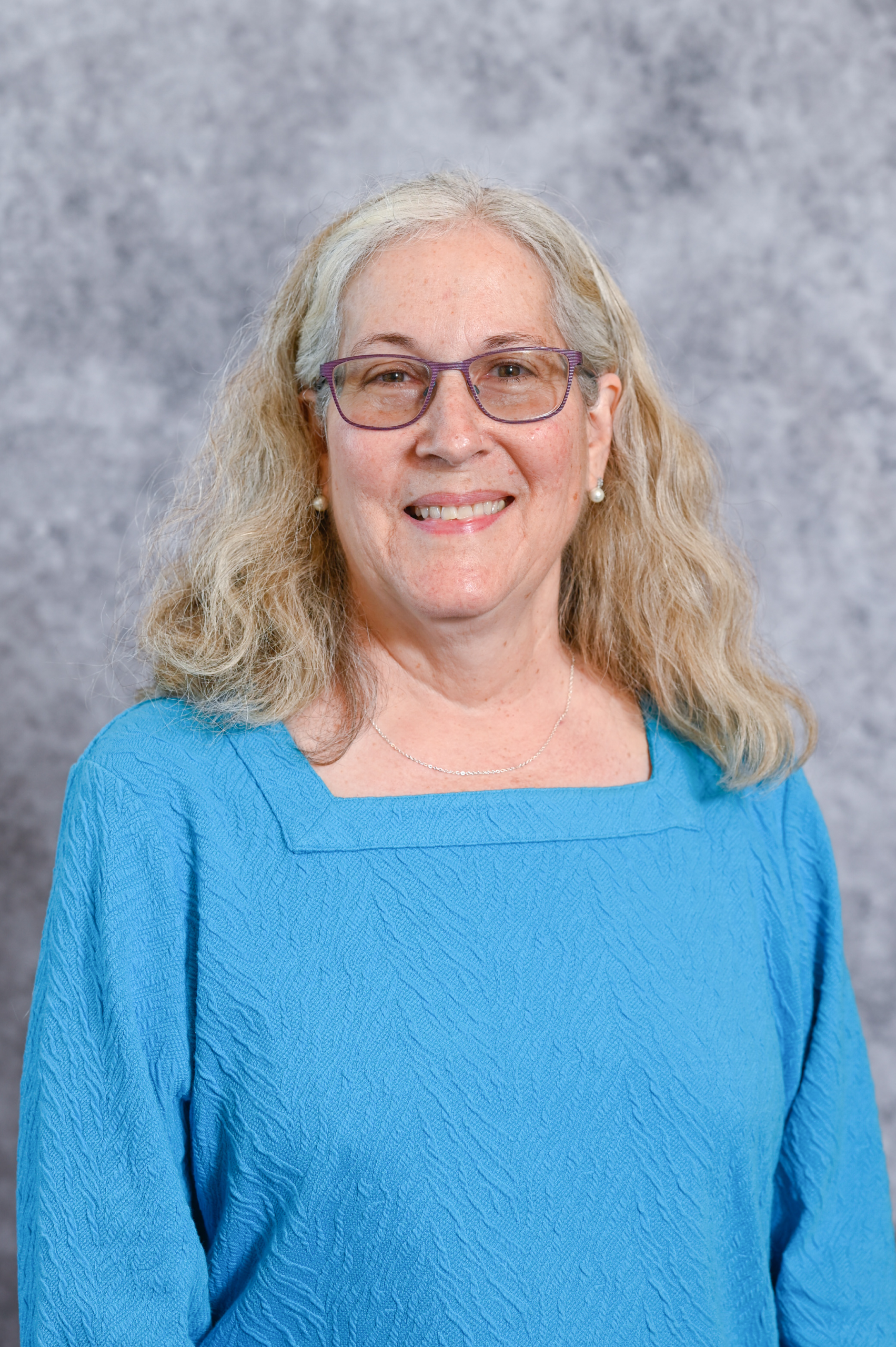 A woman with long, wavy blonde/grey hair and purple-rimmed glasses, wearing a bright blue textured blouse and smiling at the camera.