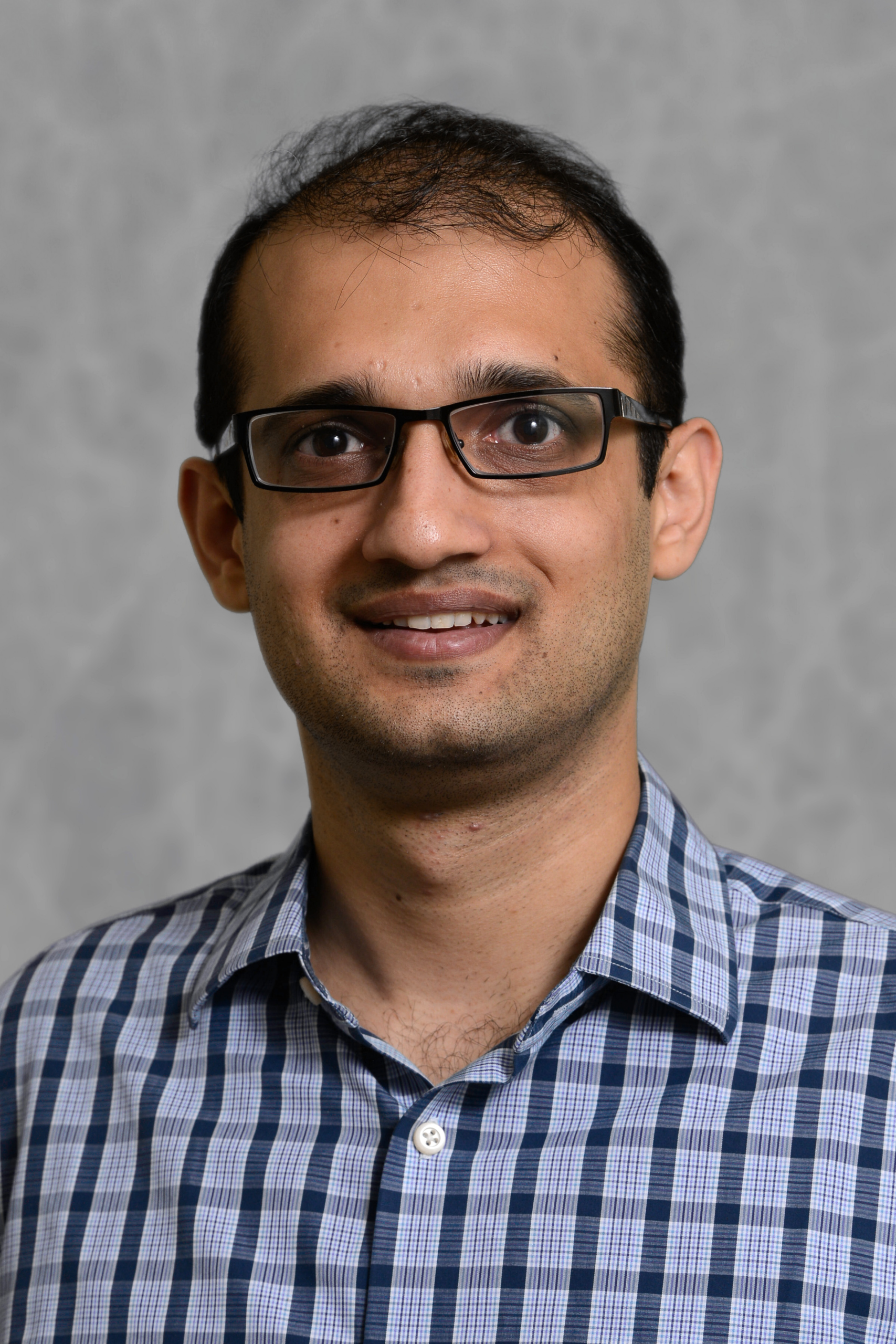 A man with short dark hair and glasses, wearing a blue and white checkered button-down shirt. He is smiling slightly against a neutral, grey textured background.