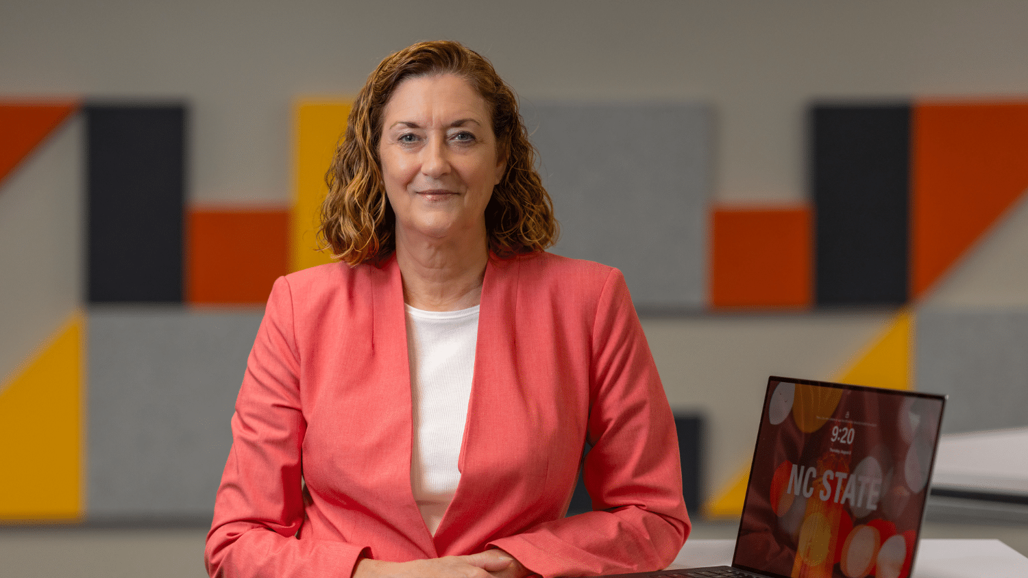 A photo of a professional woman wearing a coral blazer, sitting in an indoor space with a laptop by her side
